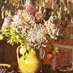 Composizione su anfora, opera di Michela Pozzato, con crisantemi, dalie, mele selvatiche, melograni e fiori secchi di aglio ornamentale e Chasmanthium.