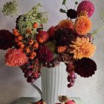 Composizione su caraffa con dalie, ammi, cinorrodi di rosa, grappoli d’uva, hydrangea essiccata, melograni e ricci di castagne.